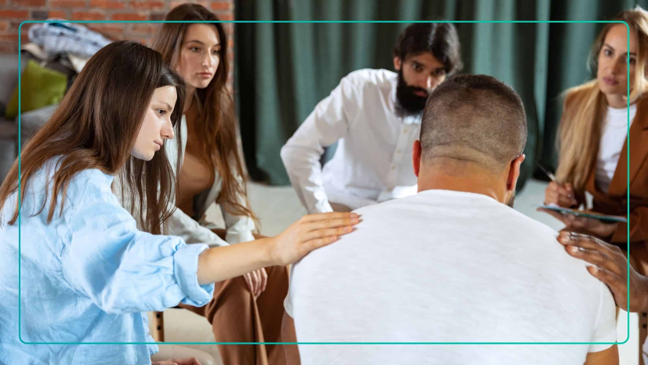 This photograph captures a small group of people seated in a circle during a group therapy session, with one participant placing a hand on another's shoulder in a gesture of support. Such sessions are often used to foster peer connection, encourage open dialogue, and provide emotional and practical assistance to members.